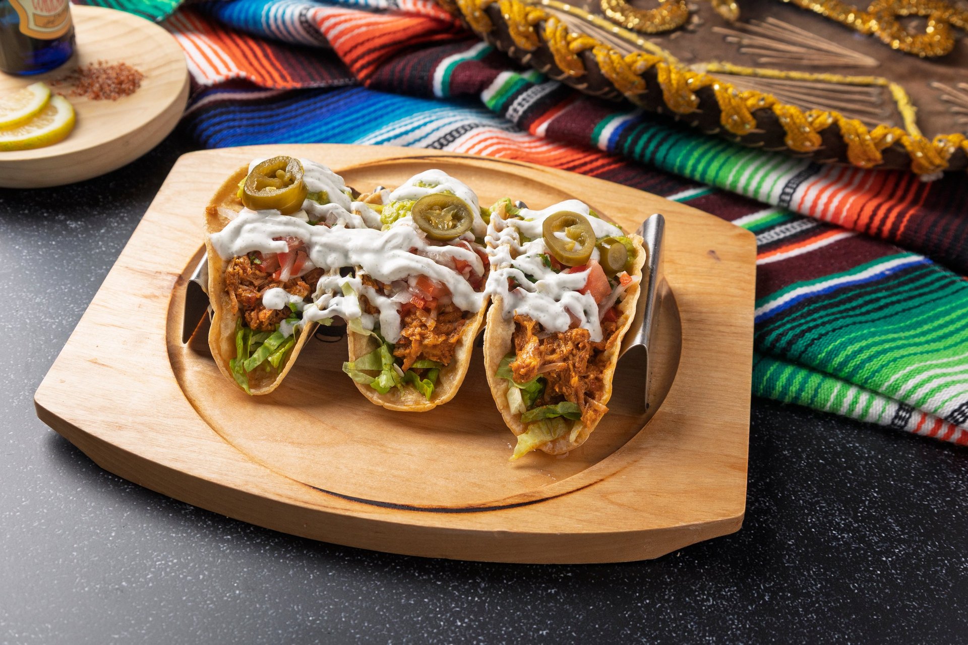 Three chicken tacos topped with jalapeños, lettuce, tomato, sour cream, and cheese served on a wooden tray with a traditional Mexican sombrero and serape
